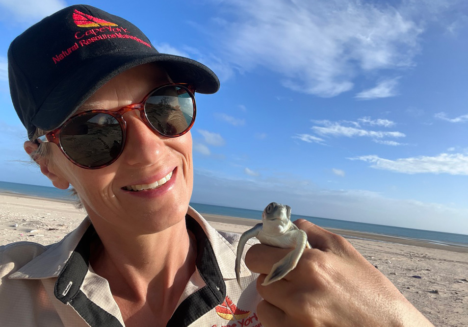 Woman in a hat and sunglasses holding a baby turtle on a beach.