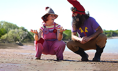 Image of two people crouching down on sand, with trees and some water in the background