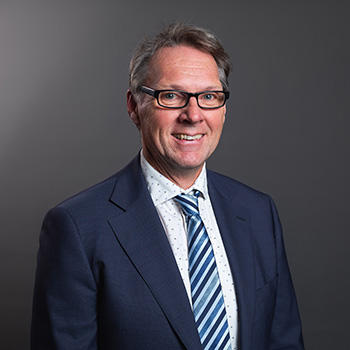 Image of Dr Bertie Henneke - a person standing in front of a grey background; they are smiling and have short hair and glasses; they're wearing a dark blue blazer; white shirt and a blue, black and white striped tie.