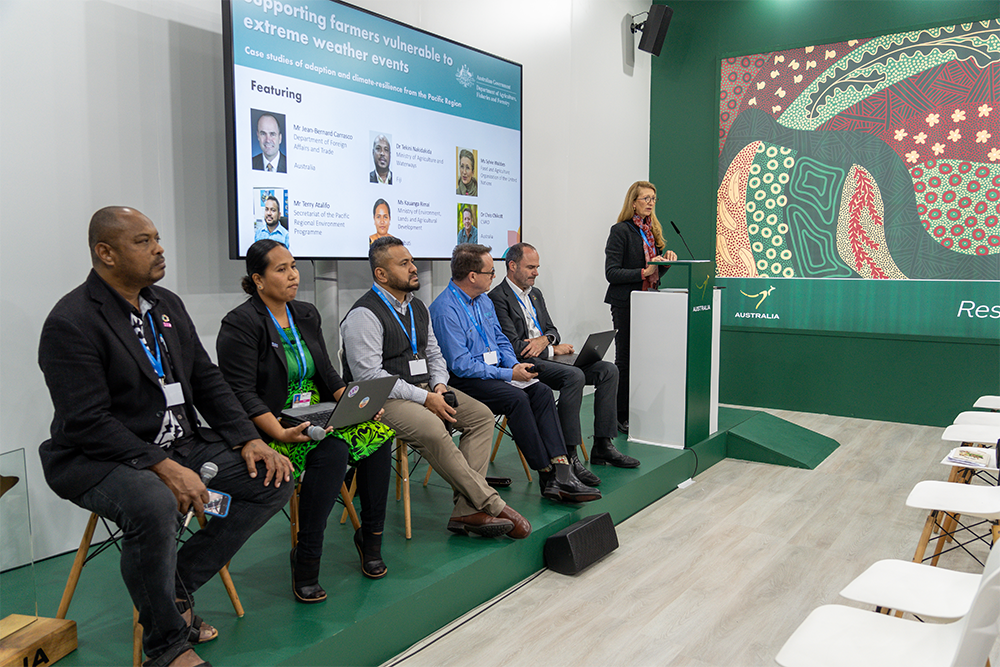 Image of 5 people sitting on a stage and another person at a podium delivering a presentation