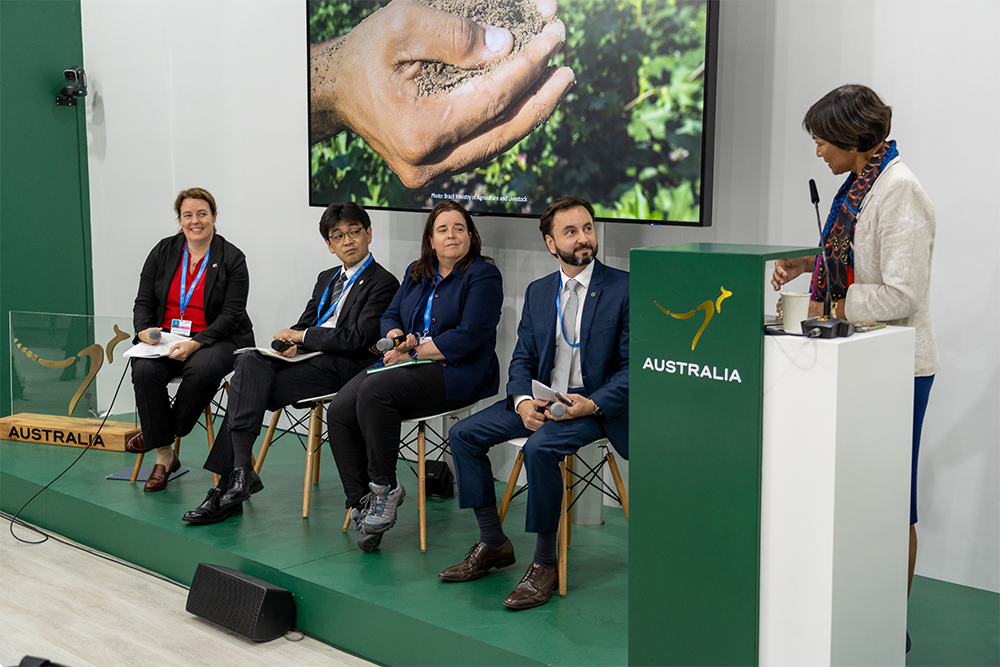 Image of 4 people sitting on a stage and another person at a podium delivering a presentation