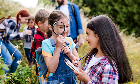 Image of several young people standing in a field, including two in the foreground, one with a large magnifying glass, another with a notepad.