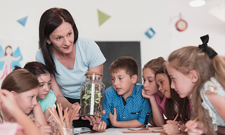 Image of a teacher standing at a table, surrounded by students; they are looking at a plant inside a glass bottle