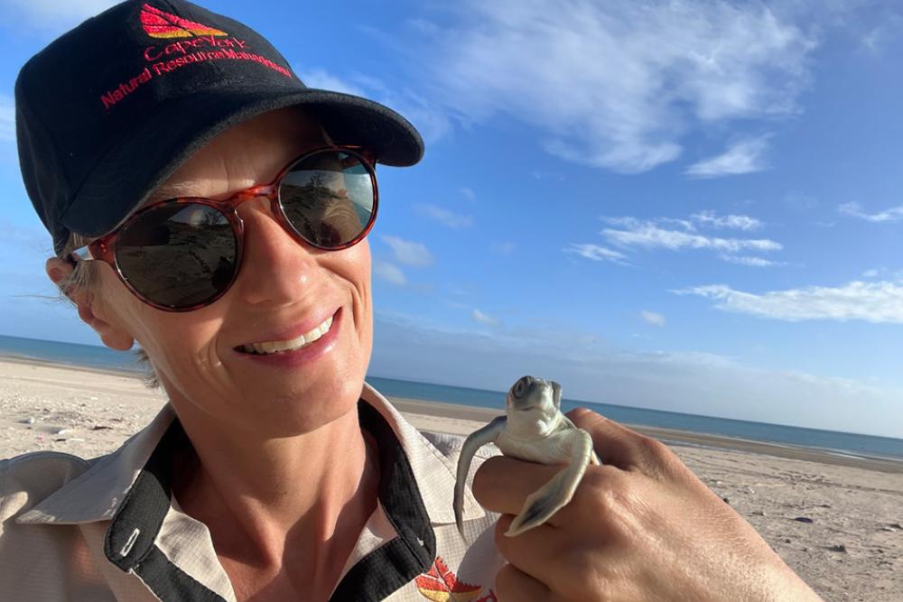 Woman in a hat and sunglasses holding a baby turtle on a beach.