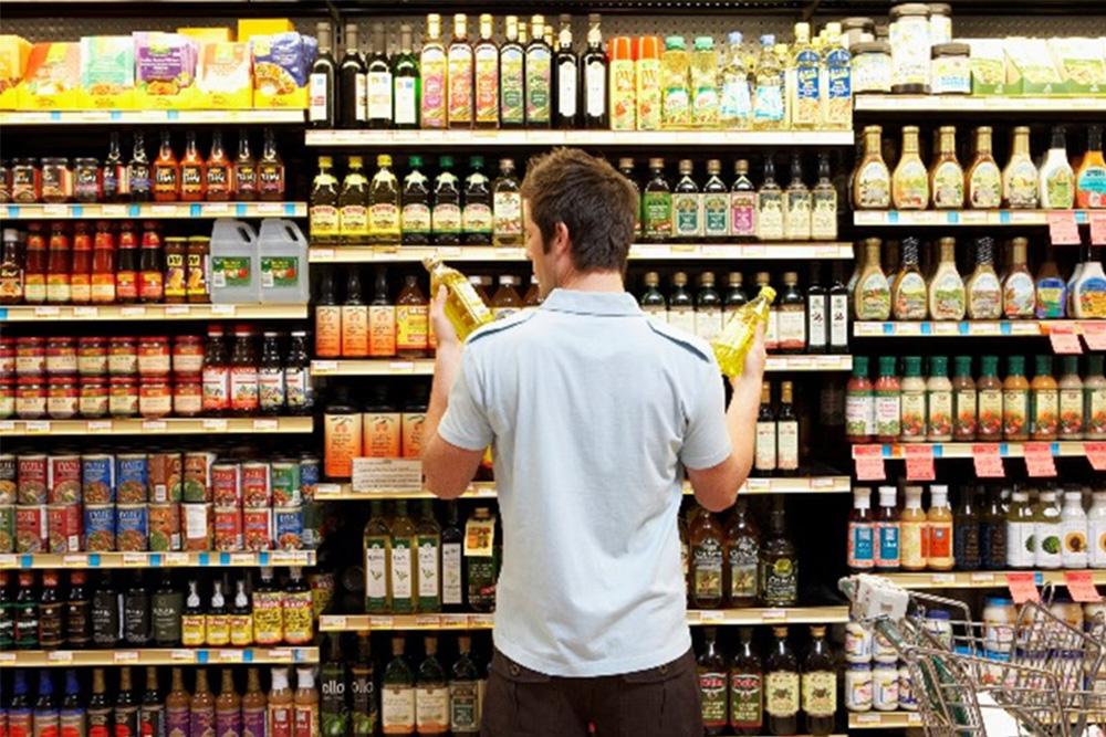 Image of a person standing in front of a shelf in a supermarket, holding up different bottles and comparing them with one another