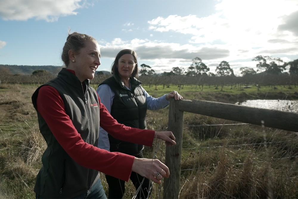 Image of two people standing at a fence in the middle of a field, with a damn and pasture in the background